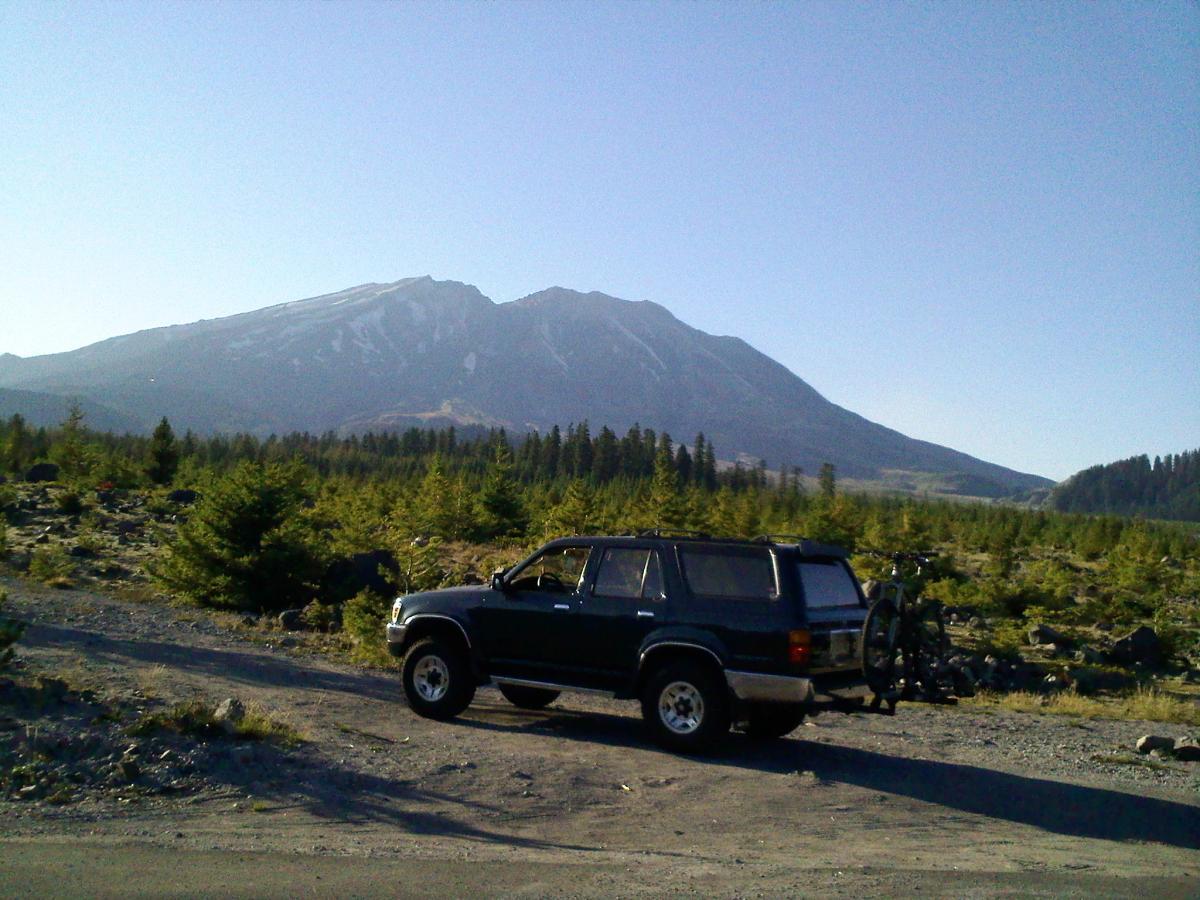 A dark green SUV parked on a rocky dirt road, with a mountain in the background. The mountain has patches of snow at the summit and is surrounded by a forest of evergreen trees under a clear blue sky. The vehicle has a bicycle mounted on the back. Ape Canyon#234, Abraham#216d, Smith Creek#225 Trails mountain bike trail.