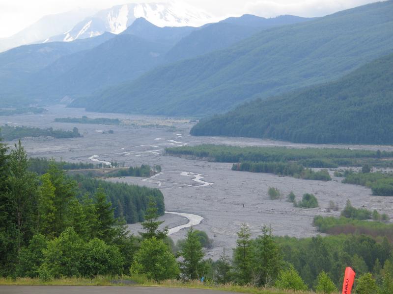 A panoramic landscape showcasing a winding river flowing through a gray, rocky valley surrounded by lush green vegetation and distant mountains. The peaks are partially covered with snow, hinting at a colder climate in the background. The scene conveys a tranquil yet rugged natural environment. Ape Canyon#234, Abraham#216d, Smith Creek#225 Trails mountain bike trail.