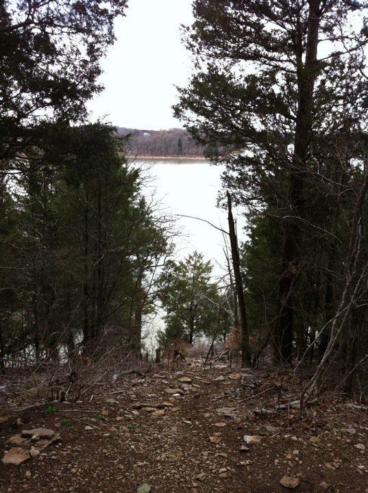 A tranquil view of a lake framed by trees, with a rocky pathway leading down towards the water. The scene is set against a cloudy sky, creating a calm and serene atmosphere. Jones Mill mountain bike trail.