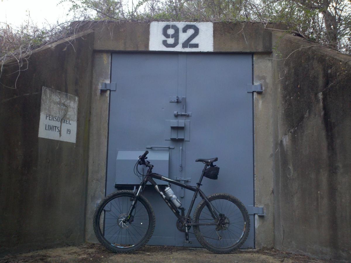 A mountain bike leaning against a large gray door marked with the number 92, situated in front of a concrete structure. To the left, a sign indicates "PERSONNEL LIMITS 19." Surrounding vegetation is visible, adding a natural element to the scene. Enterprise South mountain bike trail.