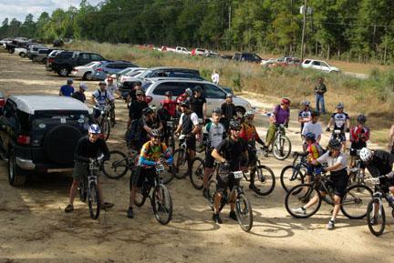 A group of mountain bikers gathered at the starting line of a race, with various bicycles and helmets, surrounded by vehicles parked in a wooded area. UWF Mountain Bike Trails mountain bike trail.