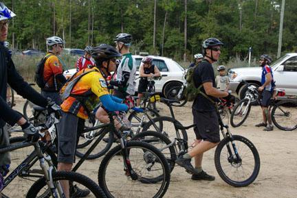 A group of mountain bikers stands near their bicycles in a forested area, preparing for a ride. They are wearing helmets and cycling gear, with various styles of bicycles lined up in front of them. A few vehicles are parked in the background. UWF Mountain Bike Trails mountain bike trail.