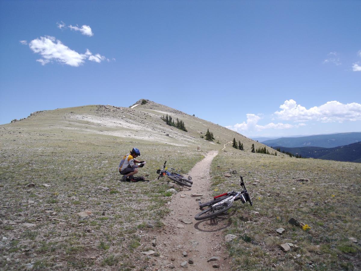A cyclist in a helmet and athletic gear crouches down on a grassy path, checking equipment near two parked mountain bikes. The landscape features rolling hills and a clear blue sky with a few clouds, adding to the serene backdrop of a mountainous area. Monarch Crest Trail mountain bike trail.