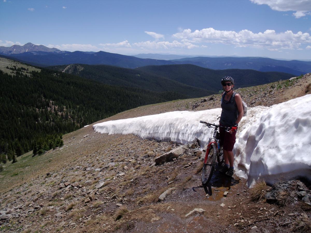 A person standing next to a snowbank on a mountain trail, holding a mountain bike. The scene features a panoramic view of rolling hills and distant mountains under a clear blue sky with some clouds. The terrain is rocky with sparse vegetation, emphasizing the high altitude. Monarch Crest Trail mountain bike trail.