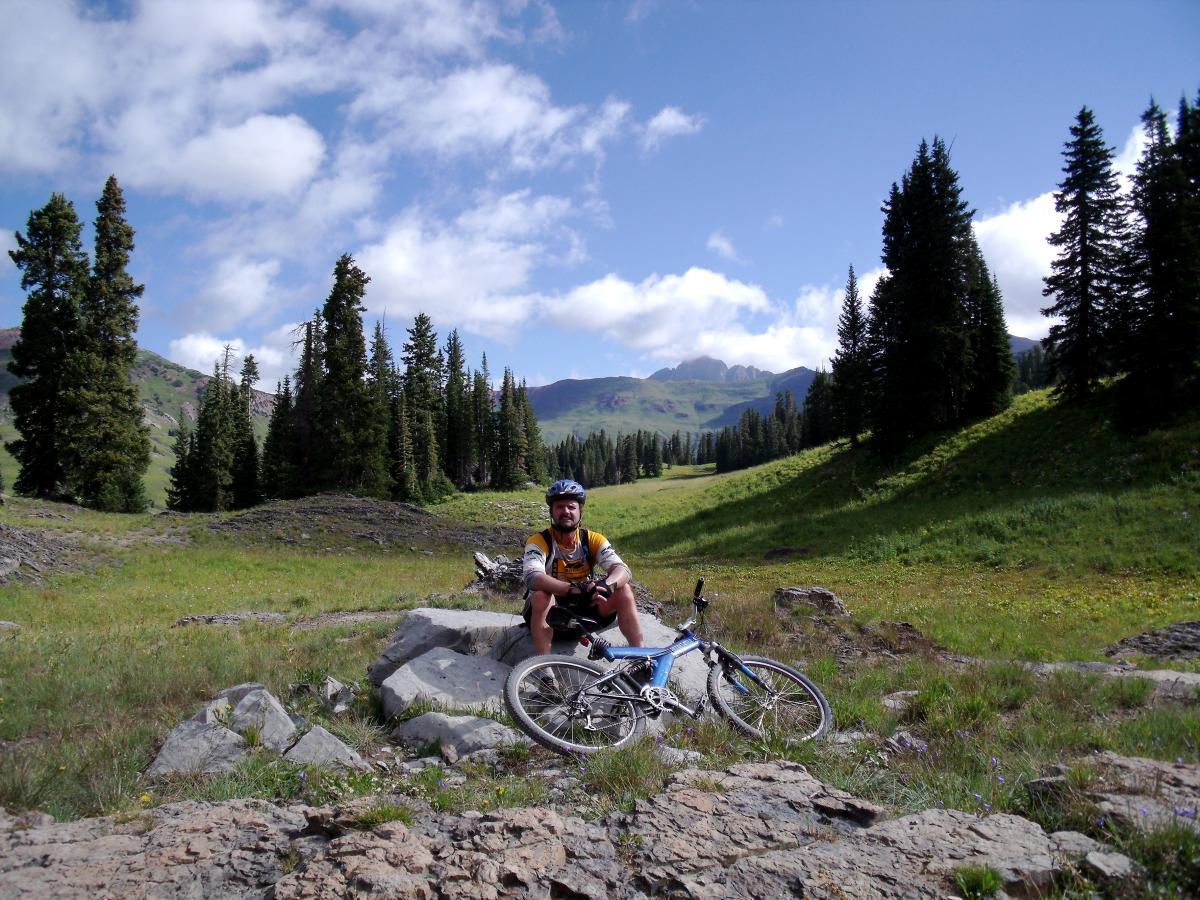 A mountain biker resting on a rock in a lush green valley, surrounded by tall pine trees and mountains under a partly cloudy sky. The biker is wearing a helmet and biking gear, with a mountain bike nearby. Trail 401 mountain bike trail.
