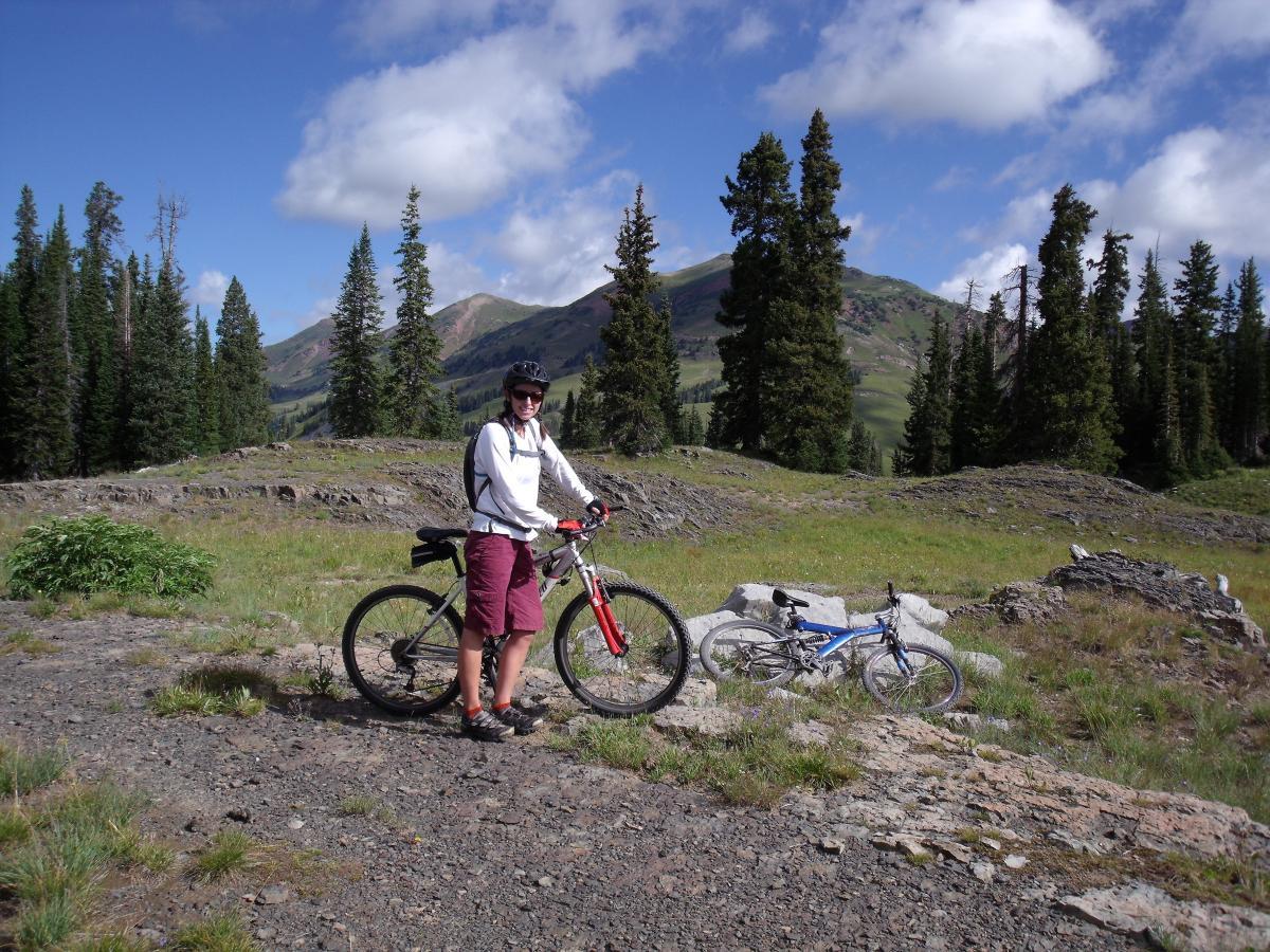 A person wearing a helmet and a long-sleeve shirt stands next to a mountain bike on a rocky terrain with trees and mountains in the background. Another bike is parked nearby, surrounded by greenery and a clear blue sky with some clouds. Trail 401 mountain bike trail.