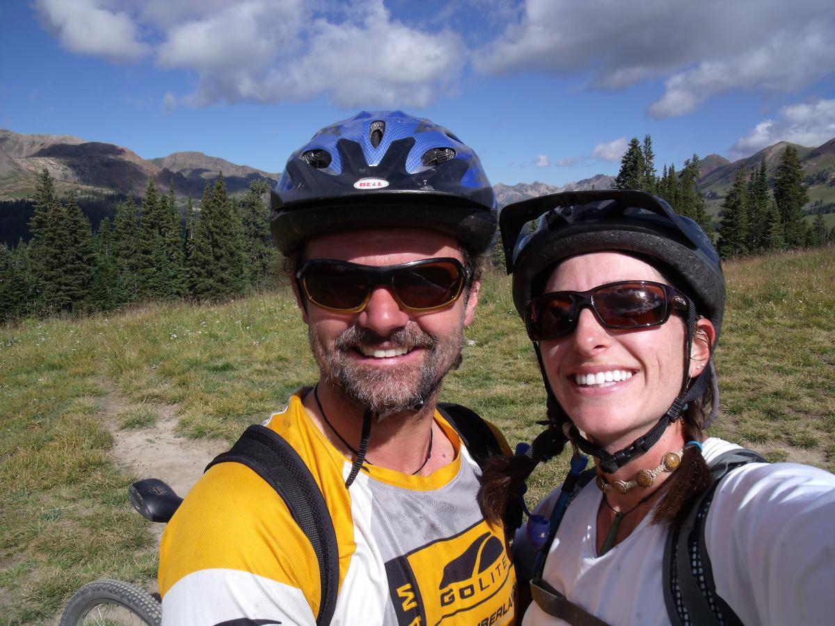 A smiling couple wearing cycling helmets and sunglasses, standing in a grassy area surrounded by trees and mountains under a partly cloudy sky. They are both dressed in activewear, ready for a biking adventure. Trail 401 mountain bike trail.