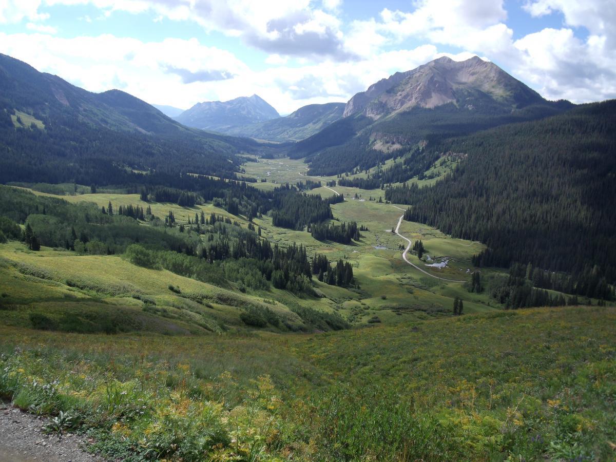 A panoramic view of a lush green valley surrounded by mountains under a partly cloudy sky. The landscape features rolling hills covered with trees, with a winding road and a stream visible in the distance. The vibrant greenery contrasts against the rugged mountain peaks, creating a serene natural setting. Trail 401 mountain bike trail.