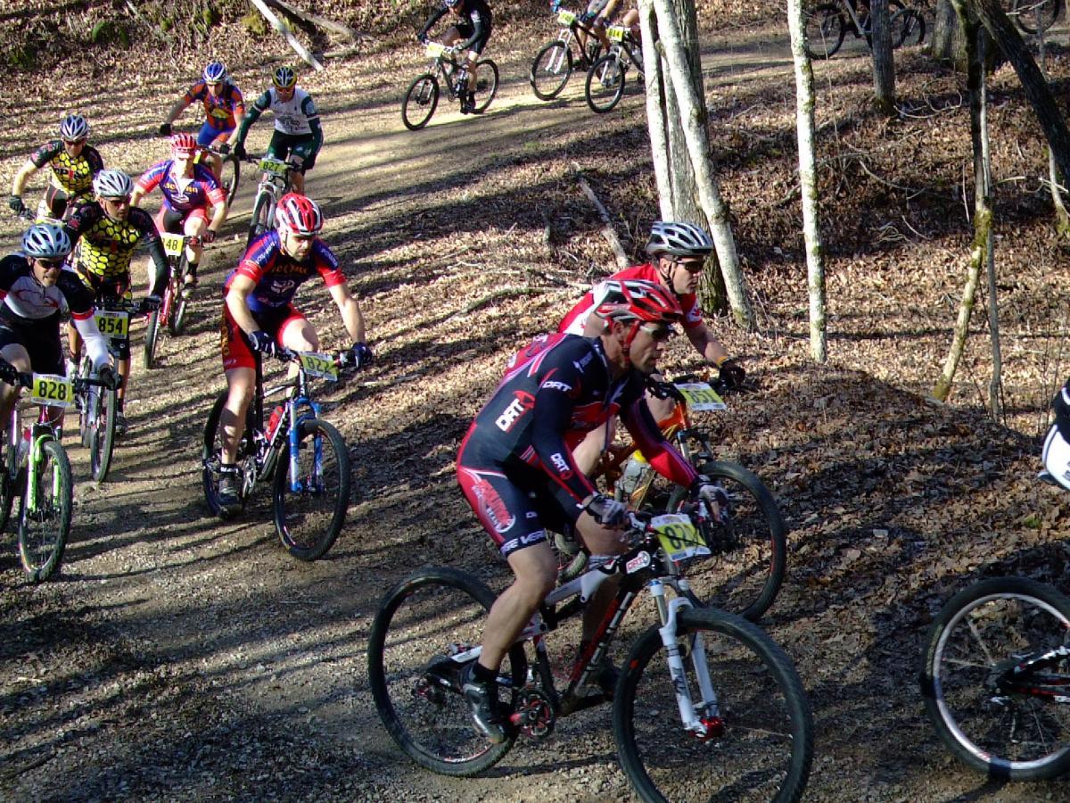 A group of cyclists competes in a mountain biking race on a dirt trail surrounded by trees. The scene captures several riders in colorful jerseys and helmets, navigating the winding path through autumn leaves. The focus is on the effort and energy of the participants as they speed along the course. Tsali Recreation Area mountain bike trail.