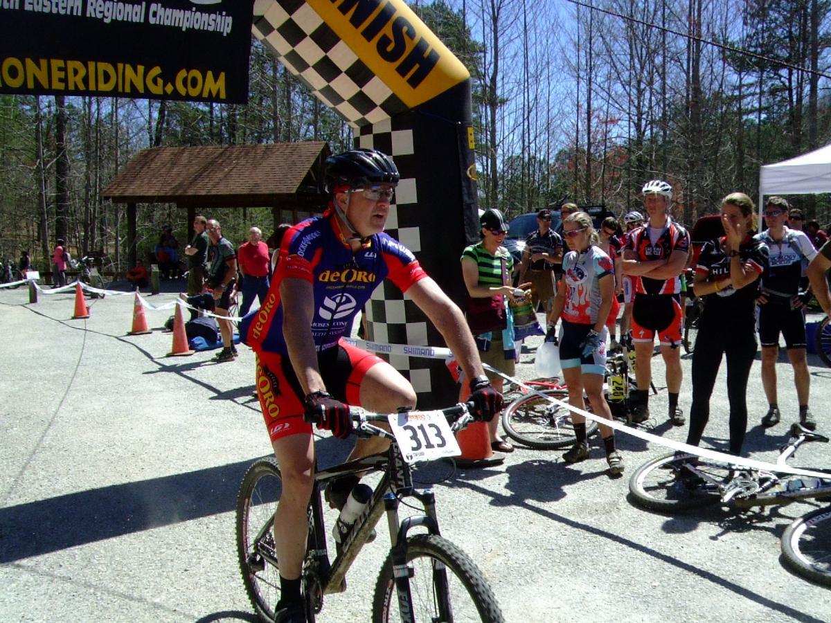 A mountain biker in colorful racing gear crosses the finish line during a regional championship event, while other competitors and spectators watch. The scene includes a large checkered archway marking the finish, with cyclists in the background and orange cones lining the path. The setting features a wooded area with trees in the distance. Tsali Recreation Area mountain bike trail.
