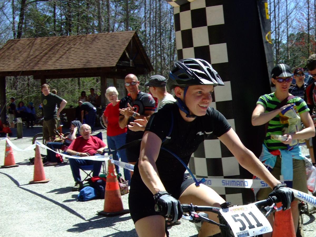 A cyclist wearing a black jersey and a helmet rides past the finish line at a bike race, smiling as they approach a cheering crowd. Spectators, including a woman in a red shirt and an older man in a red jacket, watch from the sidelines. The background features trees and a checkered banner marking the finish line. Tsali Recreation Area mountain bike trail.