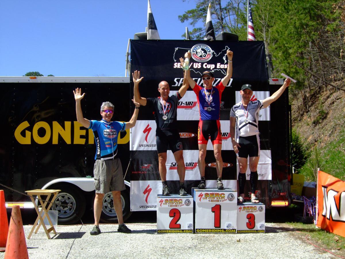 Three cyclists are standing on a podium after a competition, celebrating their achievements. The cyclist on the left, wearing a blue jersey, holds up one hand and smiles, while the middle cyclist in red holds his hands up in victory, displaying a gold medal. The cyclist on the right, dressed in white, is also smiling and holding a silver medal. The background features a trailer with "GONE RIDE" written on it and flags in the distance, under a clear blue sky. Tsali Recreation Area mountain bike trail.