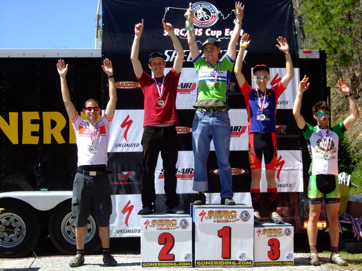 Five cyclists celebrate on a podium after a race, with the first-place winner in a green jersey holding a trophy and raising his arms in victory. The second-place finisher, in a white jersey with red accents, stands on the left with arms raised, while the third-place winner, wearing a blue jersey, is to the right of the first-place winner, also raising his arms. The backdrop features race branding and logos, with bright sunlight. Tsali Recreation Area mountain bike trail.