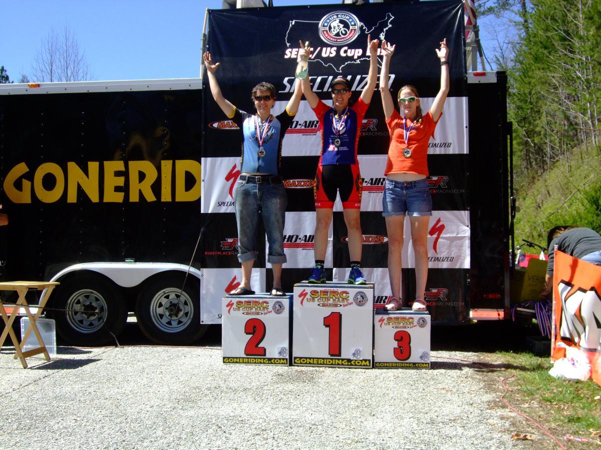 Three athletes stand on a podium celebrating their achievements at a cycling event, with the first-place winner in the middle wearing a blue jersey and holding their arms up in victory. The second-placed athlete on the left wears a silver medal and denim shorts, while the third-placed athlete on the right, wearing an orange shirt, also displays a medal. A trailer with the "Goneriding" logo and various sponsor banners serves as the backdrop, against a backdrop of trees and a clear sky. Tsali Recreation Area mountain bike trail.