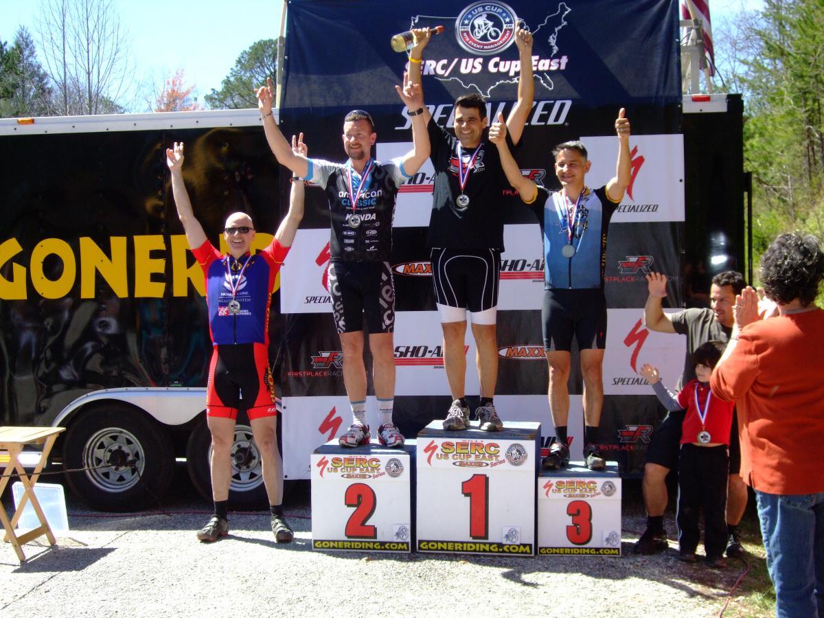 A podium at a cycling event featuring three male cyclists celebrating their achievements. The first-place winner holds a trophy, while all three athletes display medals around their necks. The background features event branding and a crowd watching. The scene is illuminated by bright sunlight. Tsali Recreation Area mountain bike trail.