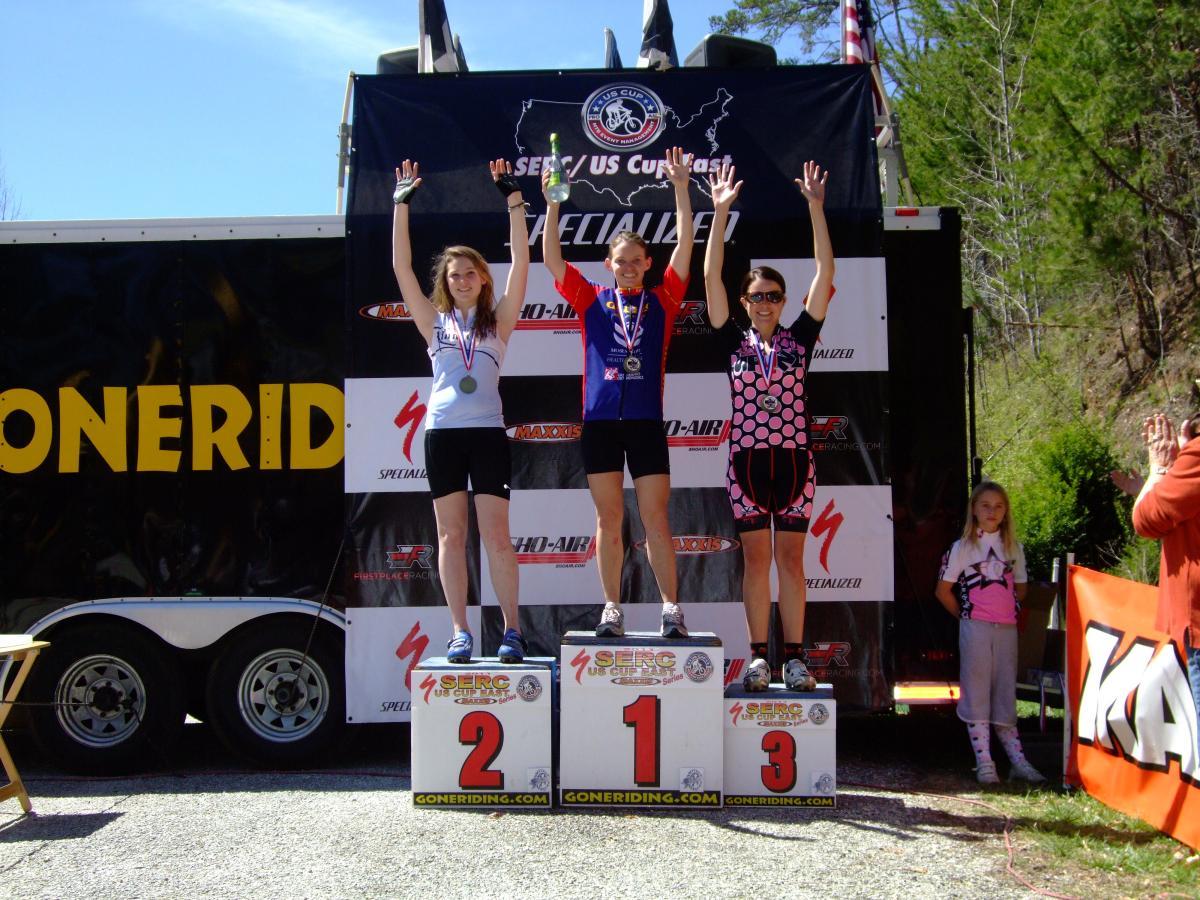 Three cyclists celebrate on a podium after a race, holding their hands up in victory. The athlete in the center, wearing a blue jersey, stands on the first place pedestal, while the athletes on either side wear medals and stand on the second and third place pedestals. A young girl is seen in the background, watching the celebration. The backdrop features sponsor logos and a clear blue sky. Tsali Recreation Area mountain bike trail.