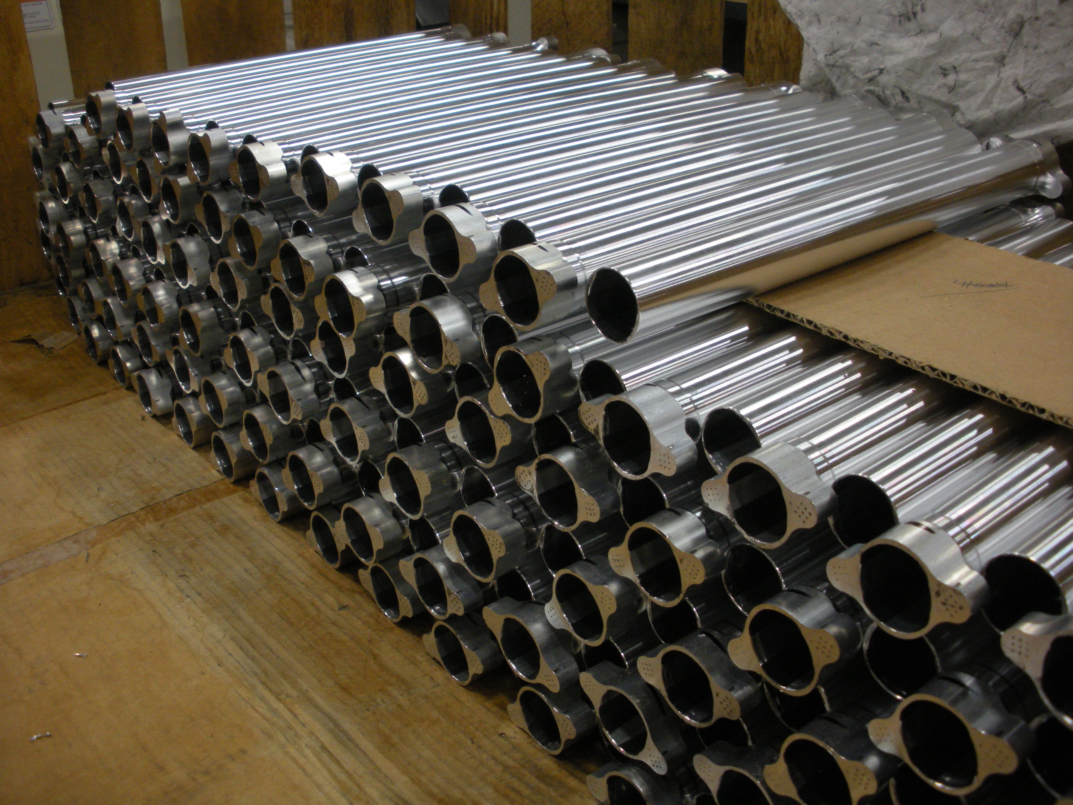 A stack of shiny metal pipes arranged in neat rows, resting on a wooden surface with some cardboard pieces visible on top. The pipes have silver exteriors and black ends, indicating they may be used for construction or industrial purposes.