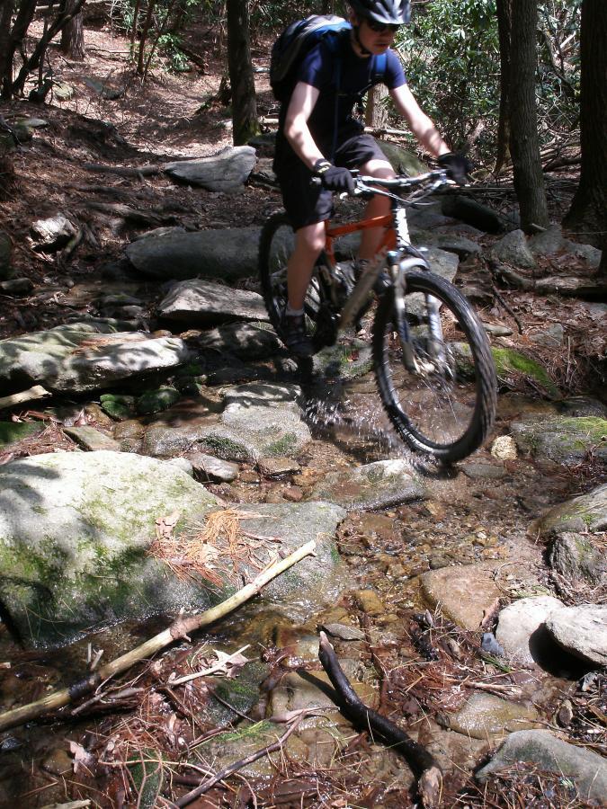 A person riding a mountain bike over rocky terrain, splashing through a shallow stream in a wooded area. Sunlight filters through the trees, highlighting the natural surroundings. The cyclist is wearing a helmet and backpack, indicating they are engaged in outdoor recreational activity. Windy Gap Trail mountain bike trail.