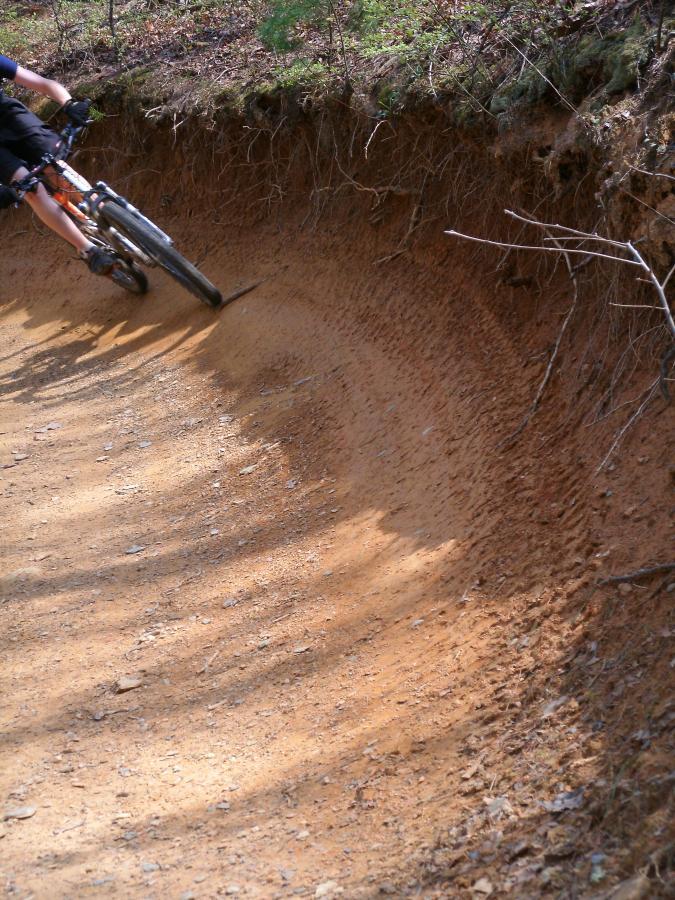 A cyclist navigating a dirt path with a curved edge, showing the textured surface of the trail. The image captures the bike leaning into the turn, emphasizing the incline of the terrain surrounded by sparse vegetation. Windy Gap Trail mountain bike trail.