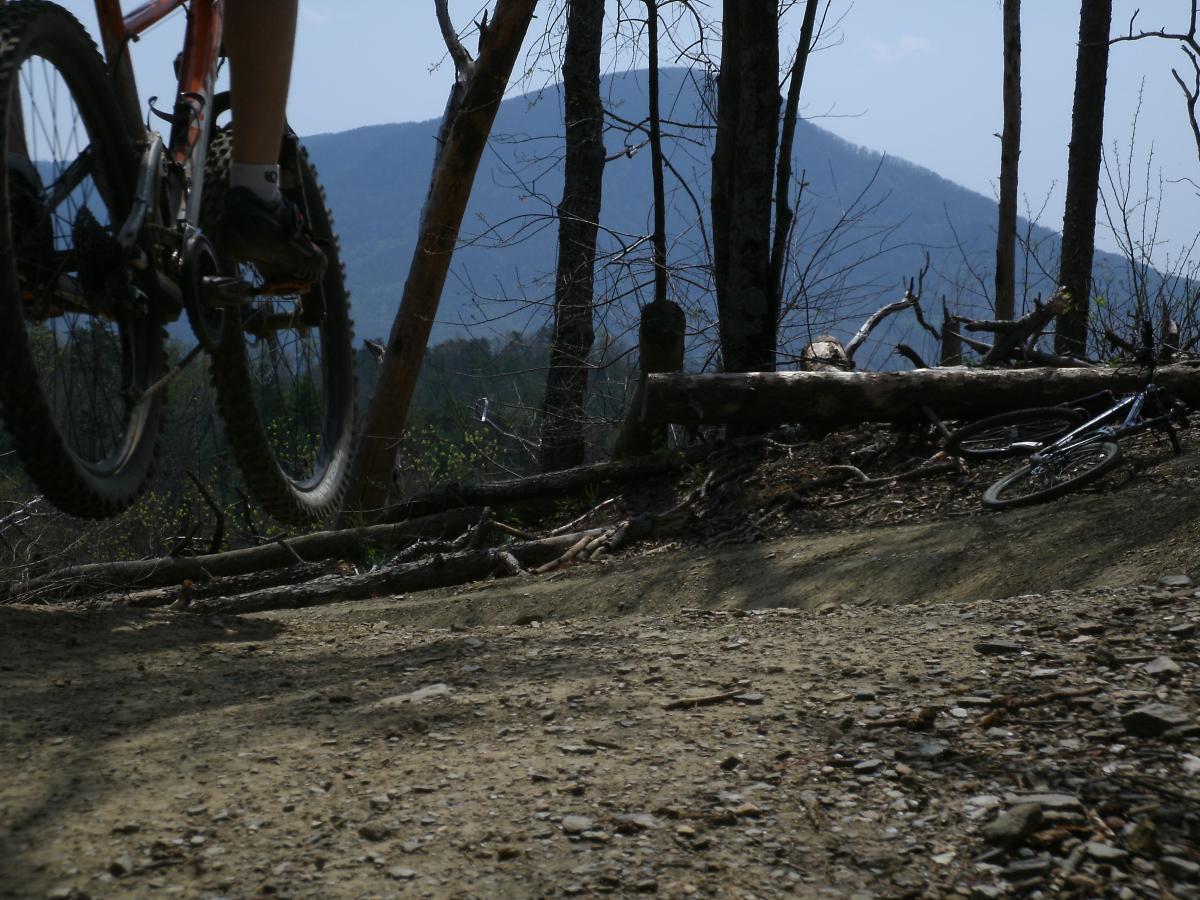 A mountain biker is captured mid-jump on a dirt trail, with a backdrop of mountains and a clear sky. Nearby, a second bicycle lies on the ground, surrounded by fallen branches and sparse vegetation. The scene conveys a sense of adventure and outdoor activity. Windy Gap Trail mountain bike trail.