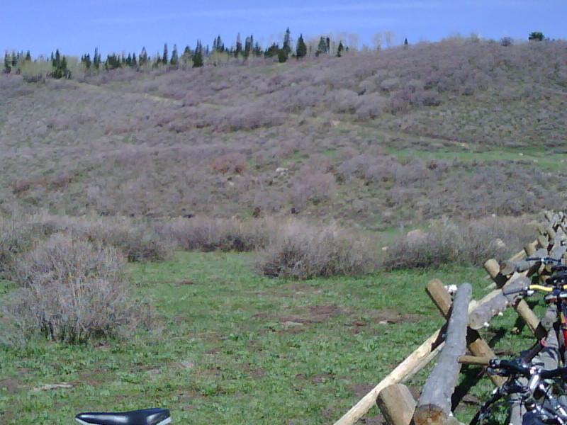 A grassy area with sparse shrubs and rolling hills in the background under a clear blue sky. In the foreground, several bicycles are parked along a wooden fence. Hazard County mountain bike trail.