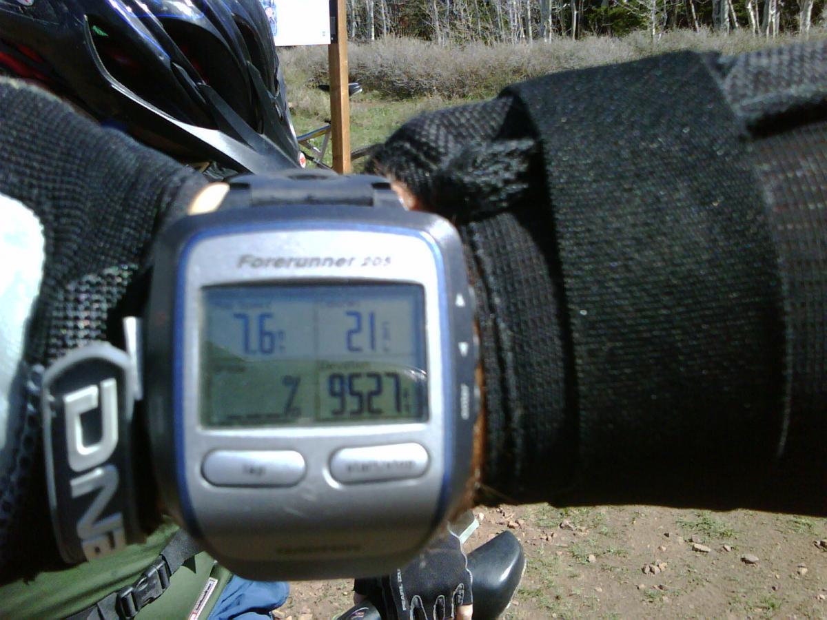 A close-up image of a wristwatch displaying statistics: a temperature of 76°F, a heart rate of 21 bpm, and an elevation of 9,521 feet. The watch is worn on a wrist covered by a black glove. In the background, several bicycle helmets are visible along with trees and a sign. Hazard County mountain bike trail.