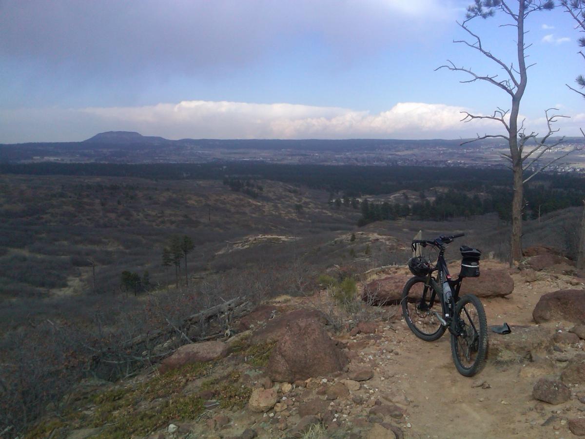 A mountain bike resting on a rocky trail overlooking a vast landscape with hills and sparse trees under a cloudy sky. The view stretches into the distance, showcasing the natural beauty of the area. Monument Preserve mountain bike trail.