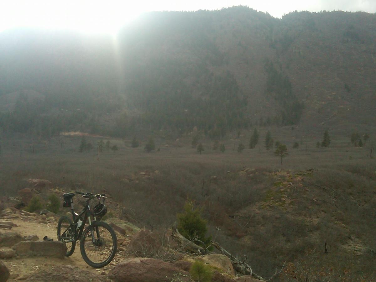 A mountain bike resting on a rocky trail with a scenic view of a vast, slightly cloudy landscape featuring hills and sparse trees under a soft, diffused sunlight. Monument Preserve mountain bike trail.