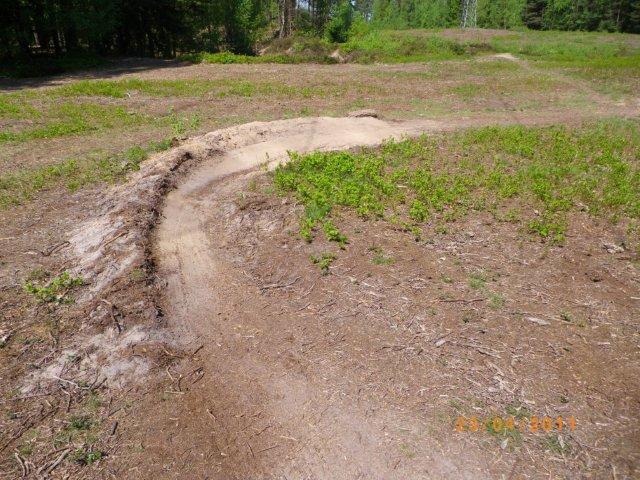 A winding dirt trail curving through a clearing, surrounded by patches of green vegetation and small shrubs, with trees visible in the background. Tiergarten mountain bike trail.