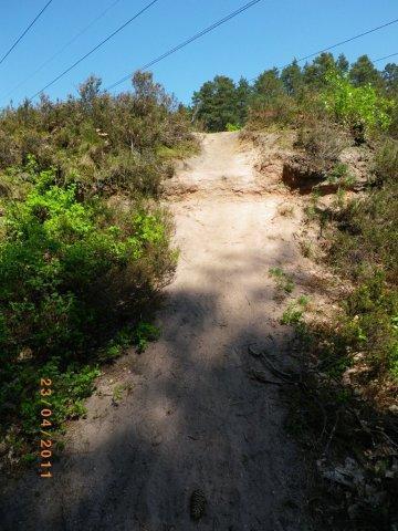 A sandy path leading uphill, surrounded by green vegetation and shrubs under a clear blue sky. Power lines are visible in the background, and the scene is bathed in bright sunlight. The date "23.04.2011" is faintly visible in the lower part of the image. Tiergarten mountain bike trail.