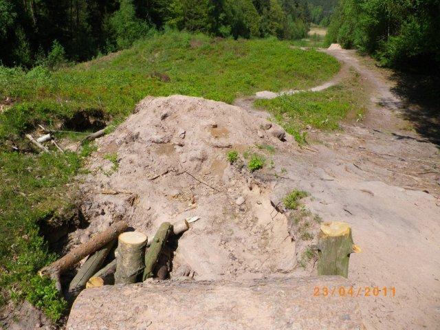 A dirt mound with exposed roots and logs sits on a forested landscape, with a visible path winding through the greenery in the background. The soil is light-colored, and patches of grass and small plants grow around the mound. The date at the bottom reads 23/04/2011. Tiergarten mountain bike trail.