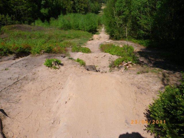 A dirt path through a sandy area, surrounded by green vegetation and trees in the background. The path is uneven, showing signs of erosion and leading into a more forested area. The image was captured on July 23, 2011. Tiergarten mountain bike trail.