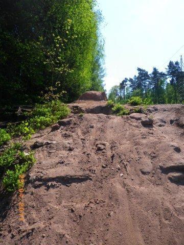 A steep, sandy incline leading up to a rocky area, surrounded by lush green trees under a clear blue sky. Tiergarten mountain bike trail.