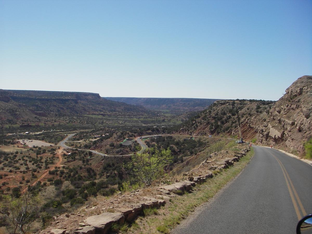 A scenic view of a winding road along a canyon, surrounded by rocky cliffs and green vegetation. The sky is clear and blue, with a distant view of layered hills in the background. A few vehicles are parked along the roadside. Palo Duro Canyon mountain bike trail.