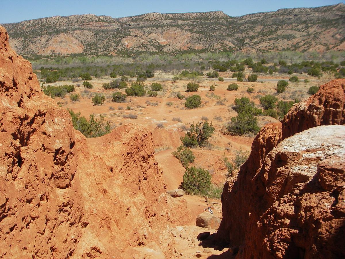 A panoramic view of a rugged, red landscape featuring rocky formations in the foreground and a wide, open plain dotted with shrubs in the background, under a clear blue sky. Palo Duro Canyon mountain bike trail.