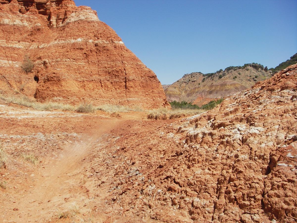 Alt text: A rocky landscape featuring reddish-brown cliffs and a pathway winding through dry terrain, with sparse grass and distant hills under a clear blue sky. Palo Duro Canyon mountain bike trail.