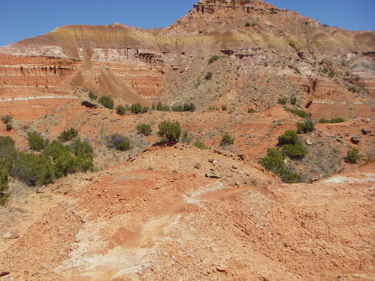 A rugged landscape featuring layered red and orange rock formations, with scattered patches of green shrubs. The scene is set under a clear blue sky, highlighting the natural erosion patterns and geological features of the terrain. Palo Duro Canyon mountain bike trail.