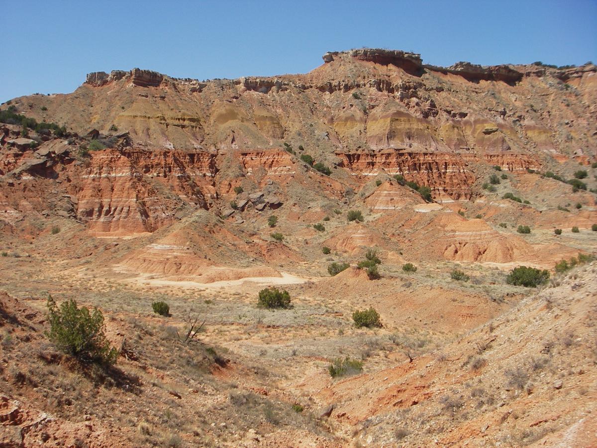 A vast desert landscape featuring rugged, multi-colored rock formations, eroded hills, and scattered vegetation under a clear blue sky. The terrain displays layers of reddish soil and rock, with gentle slopes and steep cliffs. Palo Duro Canyon mountain bike trail.