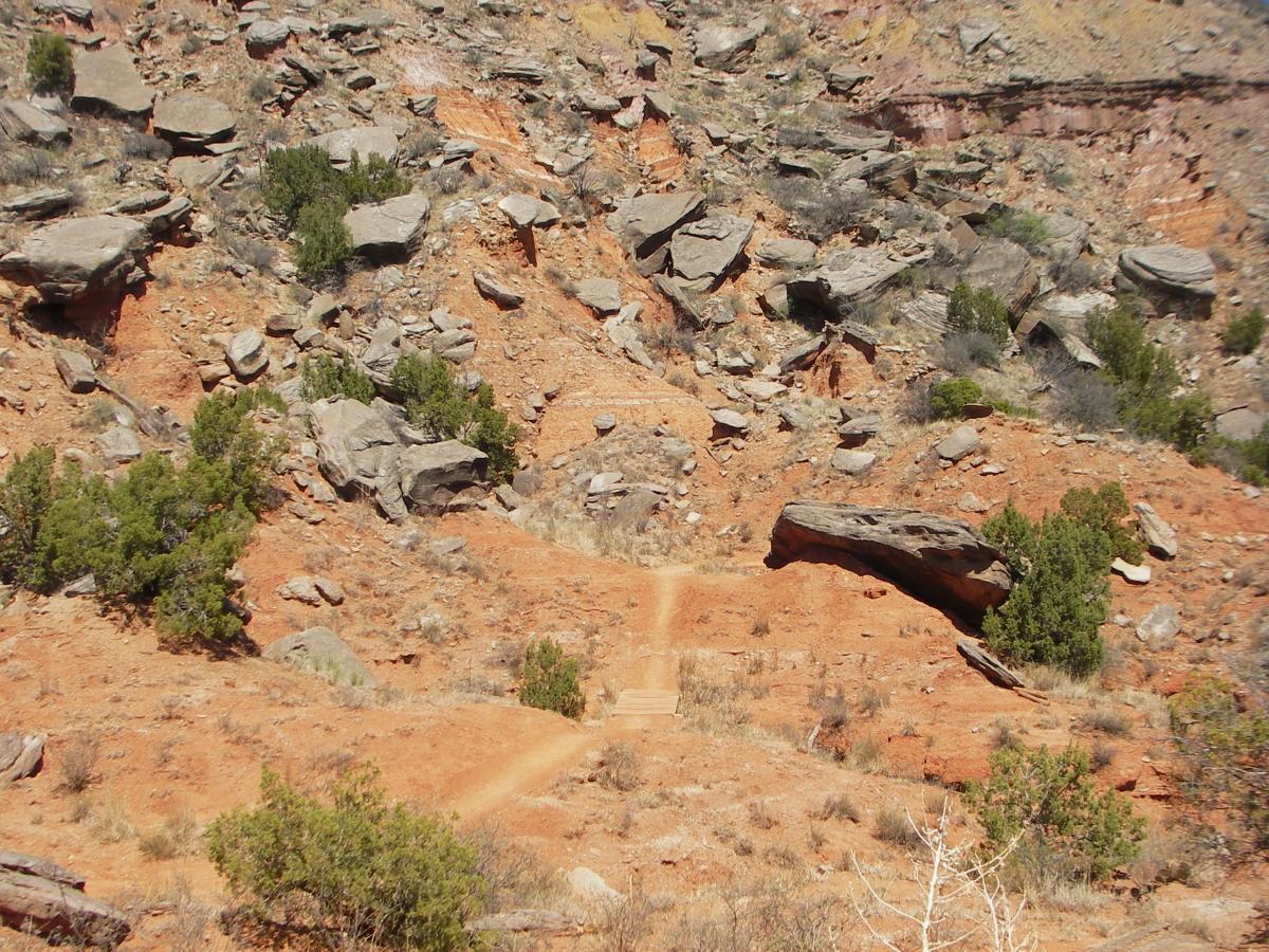 Rocky terrain with reddish-brown soil, scattered boulders, and patches of greenery. A narrow pathway winds through the landscape, leading towards a distant ridge under a clear sky. Palo Duro Canyon mountain bike trail.