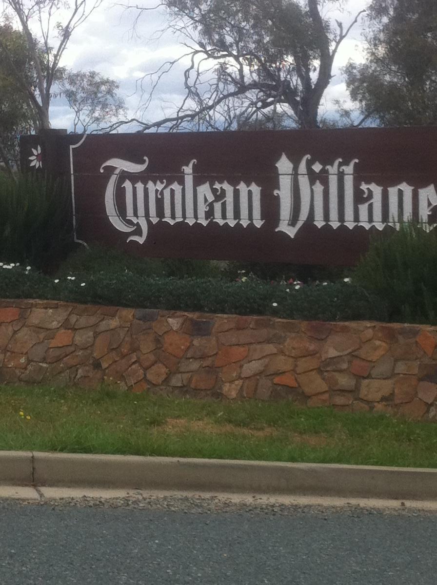 A wooden sign reading "Tyrolean Village," surrounded by greenery and a stone wall, against a backdrop of cloudy skies. Jindabyne Community Trail mountain bike trail.