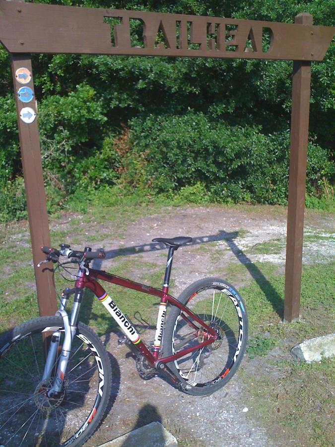 A mountain bike resting next to a wooden trailhead sign that reads "TRAILHEAD." The background features lush greenery, indicating an outdoor area for biking or hiking. The Preserve mountain bike trail.