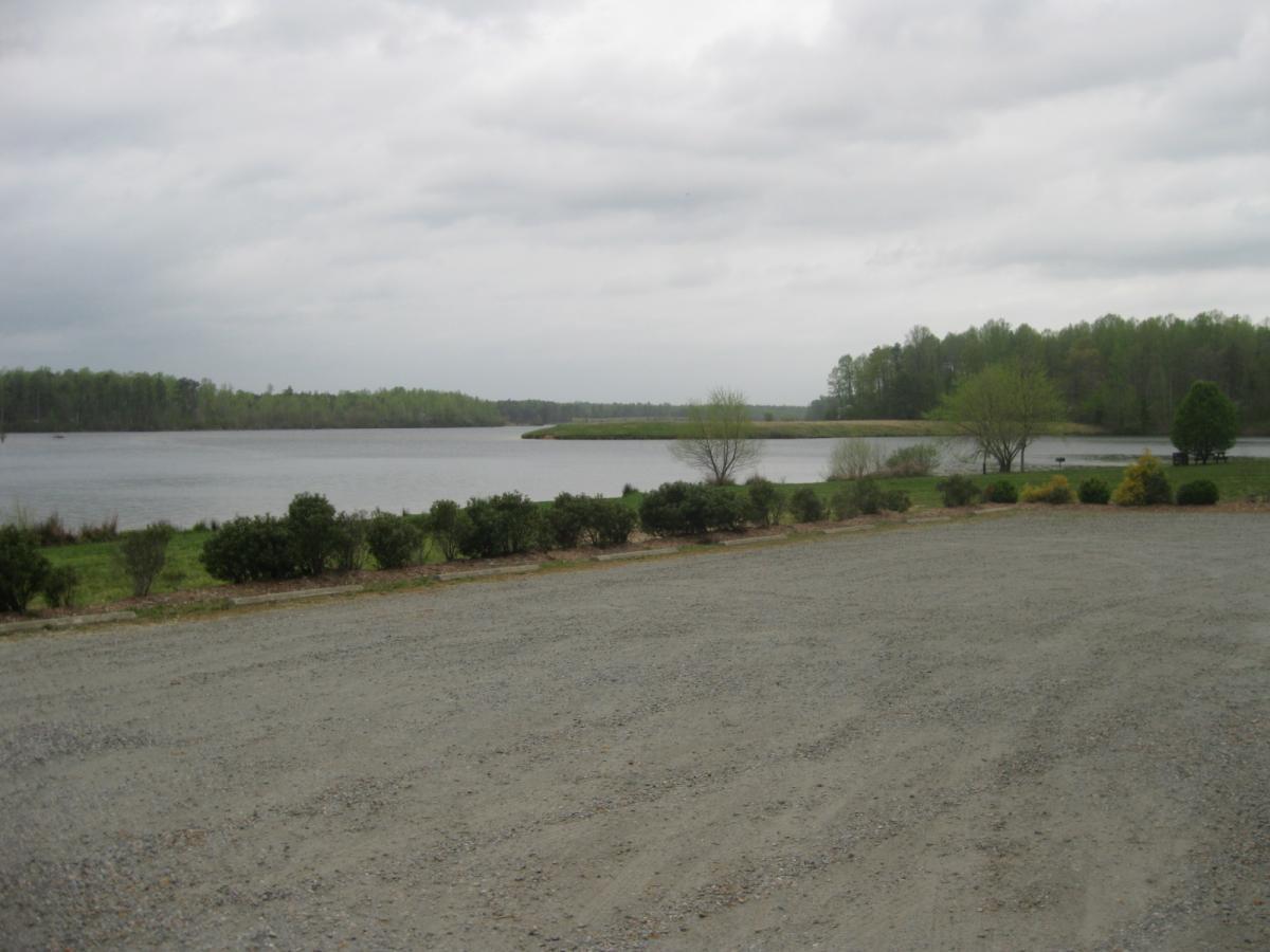 A serene landscape featuring a calm lake bordered by greenery, including trees and shrubs. The sky is overcast, creating a peaceful atmosphere. A gravel area is visible in the foreground, leading to the water's edge. Beaverdam Park mountain bike trail.