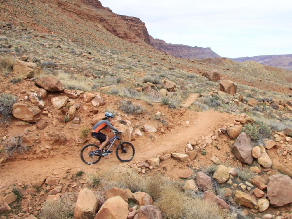 A mountain biker navigates a rocky, winding trail through a desert landscape with sparse vegetation and rugged terrain. Pipe Dream mountain bike trail.