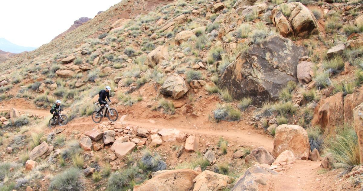 Two mountain bikers navigate a dirt trail through a rocky landscape, surrounded by sparse grass and boulders on a cloudy day. The trail winds around the rugged terrain, showcasing the beauty of outdoor cycling. Pipe Dream mountain bike trail.
