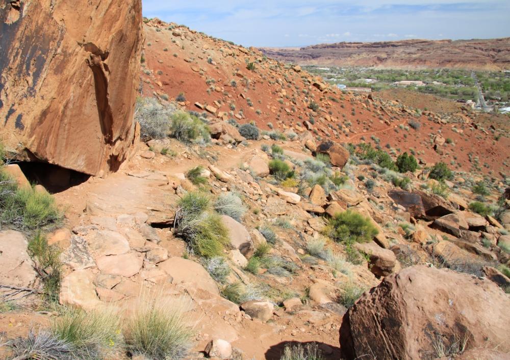 Alt text: A rocky hillside with red dirt and scattered vegetation, featuring a large boulder in the foreground and a valley with small buildings visible in the distance under a blue sky. Pipe Dream mountain bike trail.