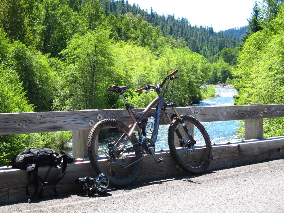 A mountain bike leaning against a wooden railing by a river, with lush green trees in the background. A backpack and helmet are placed nearby on the ground. Bright sunshine illuminates the scene, creating a vibrant outdoor atmosphere. Middle Fork Trail mountain bike trail.