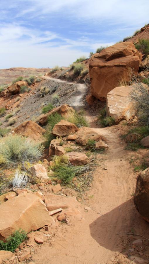 A winding dirt path through a rocky desert landscape, flanked by large boulders and sparse vegetation, under a blue sky with wispy clouds. Pipe Dream mountain bike trail.