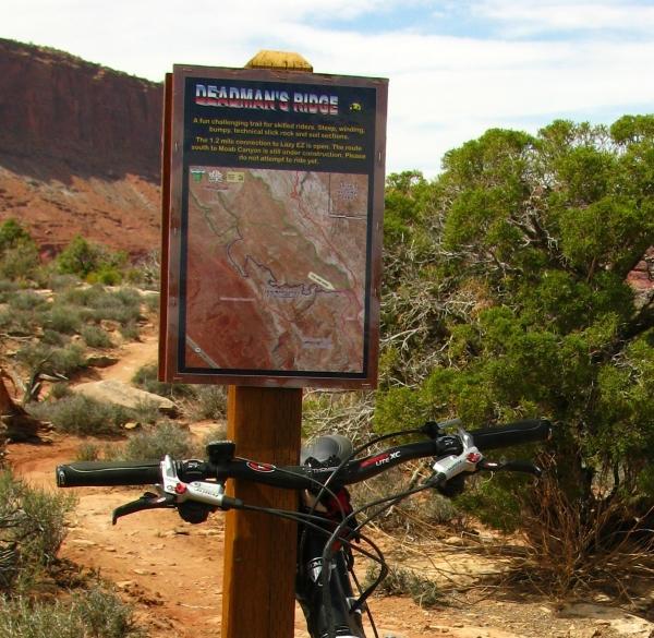 A mountain bike is parked in front of a trail sign for "Dearman's Ridge," which includes a map and descriptions of the trail. The scene features rocky terrain and shrubs, with a backdrop of red rock formations under a partly cloudy sky. Deadman's Ridge mountain bike trail.