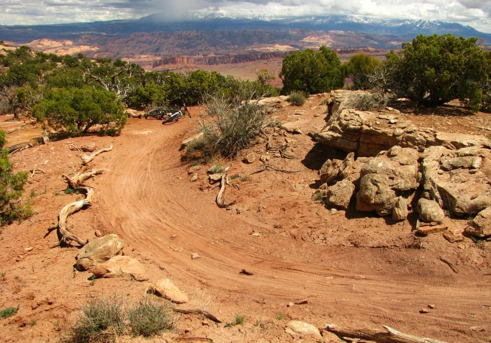 A winding dirt trail surrounded by shrubs and rocks, with a view of distant mountains under a partly cloudy sky. A bicycle is parked to the side, indicating an outdoor recreational area. Dead Horse Point State Park mountain bike trail.