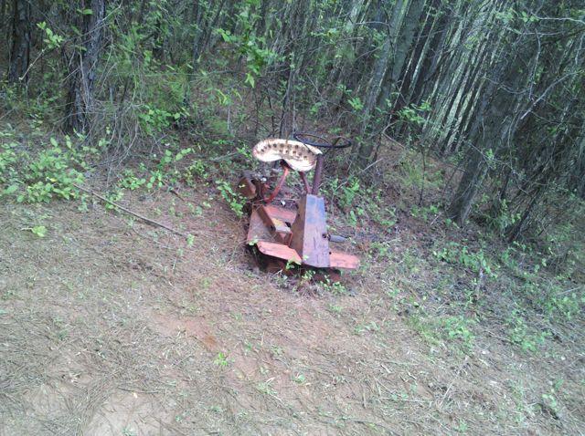 An old, rusted piece of farm equipment with a worn seat sits in a forested area, surrounded by greenery and pine needles. Taylor Randahl Memorial Mountain Bike Trails At Olde Rope Mill Park mountain bike trail.
